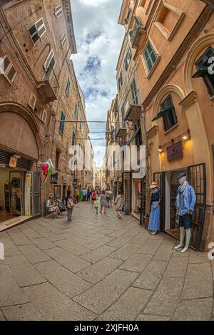 ALGHERO, ITALIA - 3 LUGLIO 2024: Pittoresca strada medievale tipica sarda con piccoli ristoranti tradizionali e negozi di souvenir. Foto Stock