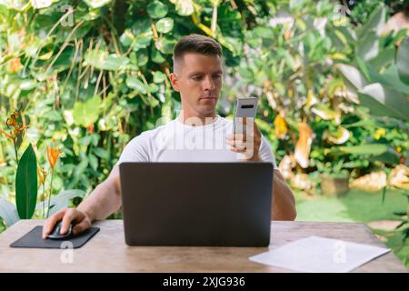 Uomo serio che tiene in mano uno smartphone e lavora su un computer portatile in un ufficio open space. Foto Stock
