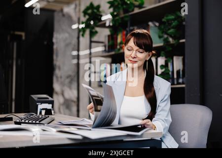 Una donna d'affari siede alla sua scrivania a rivedere i documenti. Indossa una camicetta bianca e un blazer azzurro, con i suoi lunghi capelli scuri tirati indietro in una Foto Stock