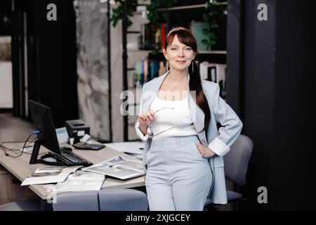 Una donna d'affari con un abito azzurro si trova in un ufficio moderno, la sua mano riposa nella tasca dei pantaloni. Tiene un paio di occhiali nell'altro Foto Stock