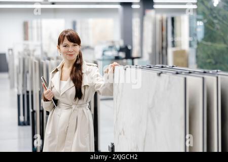 Una donna con i capelli castani e un trench beige si trova in uno showroom, guardando campioni di piastrelle. Tiene un tablet nella mano sinistra e nella mano destra Foto Stock
