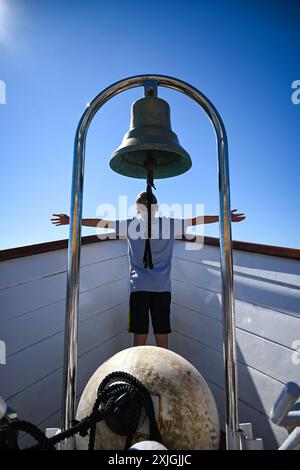 Il giovane ragazzo sta con le braccia tese sotto la campana di una nave sul ponte di una barca in una giornata di sole. Foto Stock