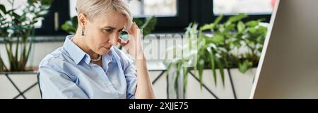 Donna d'affari di mezza età seduta alla scrivania del computer, tenendo la testa in disagio durante i sintomi della menopausa. Foto Stock