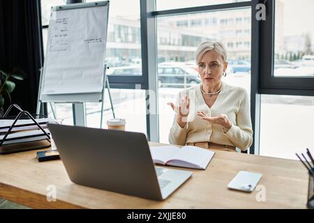 Una donna d'affari di mezza età con i capelli corti siede a un tavolo in un ufficio moderno, lavorando diligentemente sul suo notebook. Foto Stock