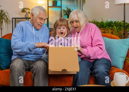 I nonni caucasici felici e stupiti e la nipotina che aprono la scatola di cartone si siedono insieme sul divano di casa. I clienti della famiglia sorridenti disimballano un buon pacco guardando all'interno un ottimo acquisto consegnato. Foto Stock