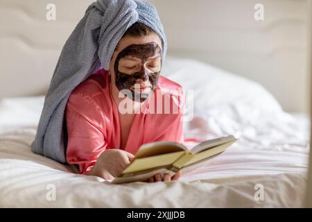 Donna con maschera nera e asciugamano sulla testa libro di lettura sul letto Foto Stock