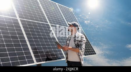 Un tecnico maschio con tablet digitale controlla il funzionamento della batteria solare. Soluzione di energia solare. Pannello solare e lavoratori sul tetto di fronte al cielo. Foto Stock