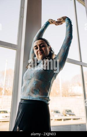 Donna sicura di sé con top in pizzo blu che si allunga vicino a una finestra Foto Stock