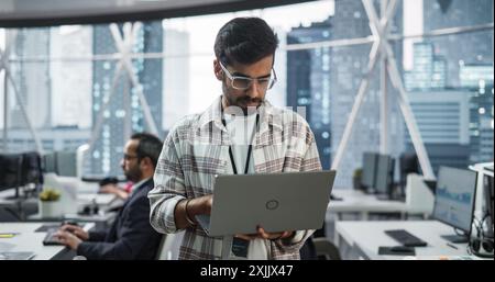 Specialista indiano professionista che utilizza un laptop mentre si trova in un ufficio vicino a una finestra con una vista sulla città. Software Data Engineer di successo Ottimizzazione del database aziendale, aggiornamento della rete di sicurezza Foto Stock