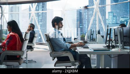 Giovani team di specialisti dell'Asia meridionale che lavorano su computer desktop in un ambiente di lavoro moderno. Sviluppatori di software indiani, manager, specialisti ambientali, sociali e di governance che lavorano in ufficio Foto Stock