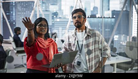 I consulenti finanziari indiani di sesso maschile e femminile utilizzano un computer portatile per discutere di una strategia di mercato azionario in un'azienda moderna. I manager dell'Asia meridionale collaborano a un progetto di ricerca e sviluppo nel settore bancario Foto Stock