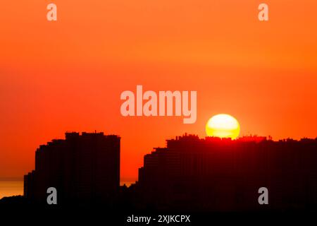 Alba nel cielo estivo sopra la città. Un grande sole che sorge sui tetti della città. Sagome di edifici moderni Foto Stock