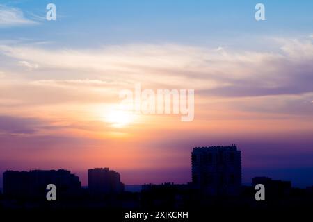 Alba in un cielo estivo nuvoloso sopra la città. Il sole attraversa le nuvole sopra i tetti degli edifici sagomati Foto Stock