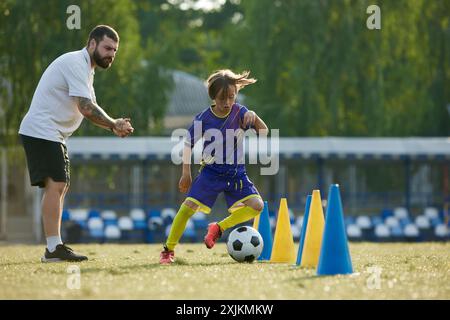 Giovane giocatore di calcio che naviga attraverso i coni, mentre l'allenatore fornisce istruzioni e supporto. Partita di football all'aperto Foto Stock