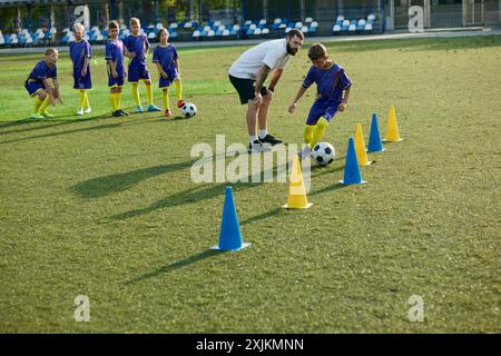 Ragazzo, bambino, giocatore di calcio che naviga attraverso i coni, mentre l'allenatore fornisce istruzioni e supporto. Partita di football all'aperto Foto Stock