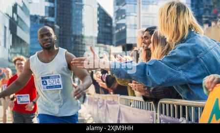 Marathon Audience applaude e applaude ai propri cari che partecipano alla gara: Black Athletic male Marathon Runner che dà un High Five a un'amica femminile che lo sostiene nel pubblico durante la corsa Foto Stock
