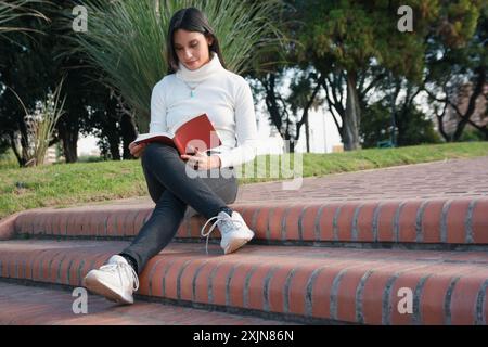 La giovane donna latina è seduta da sola al tramonto sulle scale del libro di lettura del parco, indossa un maglione bianco ed è con le gambe incrociate, è felicemente conc Foto Stock