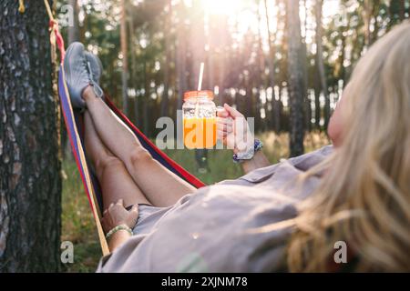 Donna che si rilassa nell'amaca nella foresta al tramonto e beve frullato o succo d'arancia Foto Stock