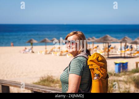 Donna turistica di mezza età che ammira la spiaggia di Praia de Alvor dopo un'escursione in Algarve, Portogallo. La donna si sta godendo una vacanza estiva attiva Foto Stock