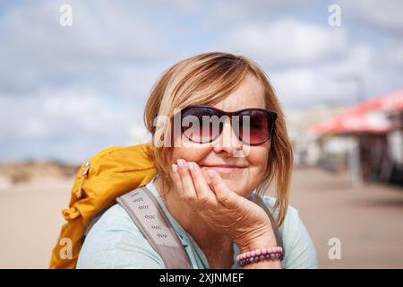 Ritratto di una donna matura e felice sorridente con occhiali da sole e zaino che si rilassa in spiaggia durante un'escursione lungo la costa dell'Algarve, Portogallo Foto Stock