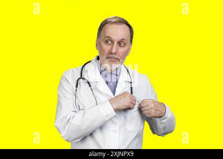Un medico anziano con stetoscopio mette i suoi soldi in una tasca per cappotto. Isolato su giallo. Foto Stock