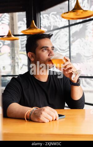 Un giovane uomo in camicia nera siede ad un tavolo da bar in legno, gustando un bicchiere di birra in un bar moderno ed elegante. L'atmosfera è caratterizzata da un'illuminazione unica e. Foto Stock