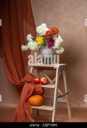 Un grande bouquet di fiori bianchi, gialli, rossi e autunnali. Molti astri in un vaso grigio su una scala di legno. Di seguito sono riportate due mele rosse, una piccola zucca. Oppure Foto Stock
