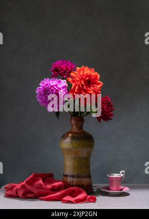 Natura morta con fiori. Dahlias rosa e rosse in vaso scuro, tessuto rosa, piccola tazza rosa, sfondo scuro Foto Stock