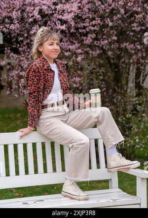 Una ragazza carina e moderna siede su una panchina del parco. Tenendo in mano una tazza di caffè. Si gode la vita e le bevande. Foto Stock