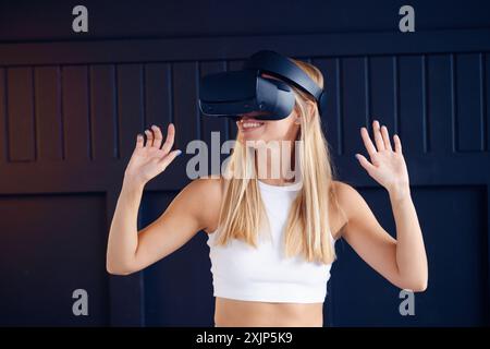 Una donna bionda in canotta bianca sorride usando il visore VR davanti al muro blu scuro, interagendo con il mondo virtuale usando le mani. Foto Stock