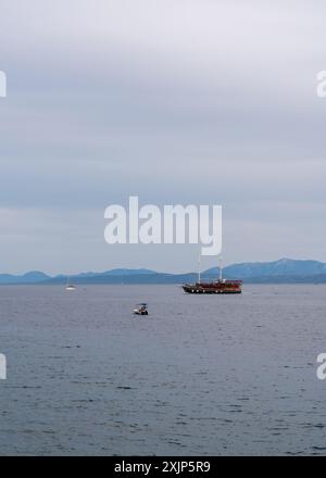 Una vecchia barca di legno con molti turisti a bordo nel Mare Adriatico. È sera. Alte montagne all'orizzonte. Piccoli yacht e barche navigano nelle vicinanze. C Foto Stock