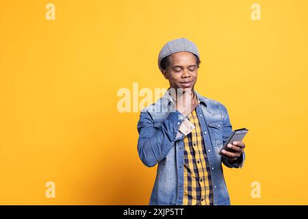 Persona matura e rilassata che pensa a una risposta ai messaggi sullo smartphone, guardando i suoi messaggi sull'app dei social media su sfondo giallo. Modello maschile sicuro che utilizza il telefono cellulare. Foto Stock