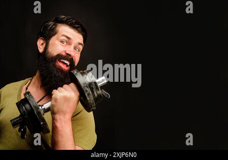 Culturista con barba atleta che si allena con manubri. Forte e bellissimo modello di fitness maschile che solleva il manubrio in palestra. Uno sportivo sorridente Foto Stock