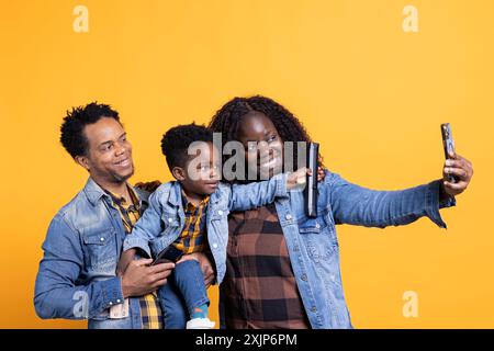 Felice famiglia afroamericana di tre persone che scattano foto in studio, posa insieme al loro bambino. Mamma e padre allegri scattano selfie sullo smartphone, creando ricordi. Foto Stock