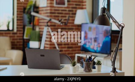 Intimo ed elegante appartamento vuoto, con TV lasciata aperta sullo sfondo e computer portatile sulla scrivania. Luminoso e moderno soggiorno con pareti in mattoni e grazioso design degli interni, primo piano Foto Stock