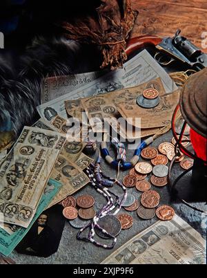 Un assortimento di monete antiche, banconote e perline giacciono su un tavolo di legno intemprato. Una pochette in pelle indossata, una pistola vintage e un vecchio stile Foto Stock