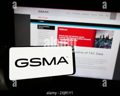 Stoccarda, Germania, 11-27-2023: Persona che detiene un cellulare con il logo dell'organizzazione del settore mobile GSM Association (GSMA) davanti all'azienda Foto Stock