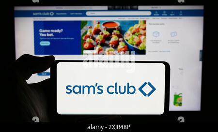 Stoccarda, Germania, 02-07-2023: Persona che detiene un cellulare con il logo della società di negozi al dettaglio Sam's Club, solo affiliata agli Stati Uniti, davanti all'azienda Foto Stock