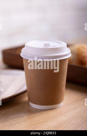 Una tazza da caffè da asporto con un coperchio bianco e un rivestimento in carta marrone, lo sfondo è leggermente sfocato, creando un'atmosfera accogliente e invitante Foto Stock