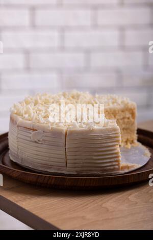 Una fetta di cremosa torta al cioccolato bianco viene servita da un piatto di legno. La torta è ricoperta da scaglie di cioccolato bianco Foto Stock