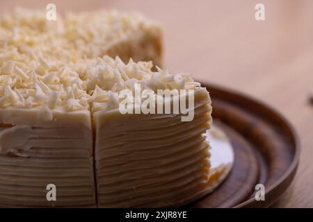 Una fetta di cremosa torta al cioccolato bianco viene servita da un piatto di legno. La torta è ricoperta da scaglie di cioccolato bianco Foto Stock