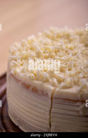 Una fetta di cremosa torta al cioccolato bianco viene servita da un piatto di legno. La torta è ricoperta da scaglie di cioccolato bianco Foto Stock