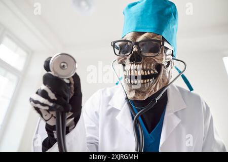 Medico che indossa la maschera scheletrica dello scull in uniforme medica bianca in piedi con stetoscopio in clinica Foto Stock