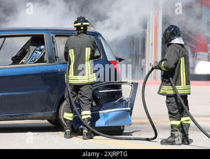 i vigili del fuoco in uniforme con caschi estinguono l'incendio dell'auto distrutta dopo l'incidente con il grande tubo dell'idrante antincendio sulla strada Foto Stock