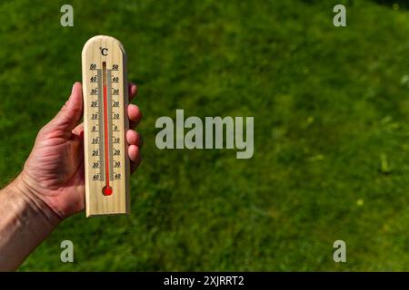 Un uomo tiene in mano un termometro di legno in giardino che indica una temperatura molto elevata - protezione contro il calore Foto Stock