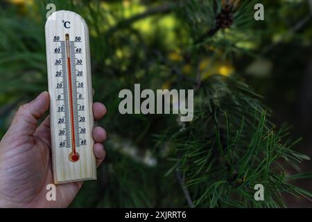 Un uomo tiene in mano un termometro di legno in giardino che indica una temperatura molto elevata - protezione contro il calore Foto Stock