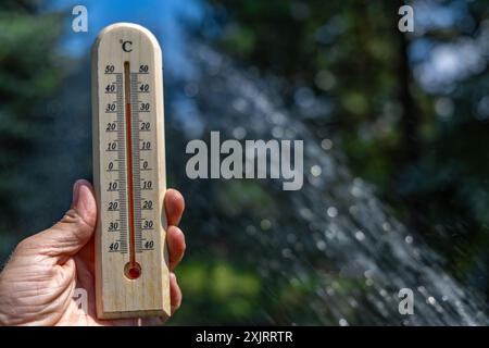 Un uomo tiene in mano un termometro di legno in giardino che indica una temperatura molto elevata - protezione contro il calore Foto Stock