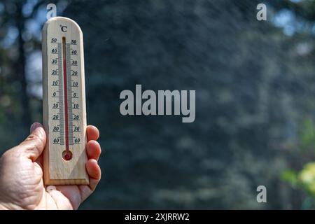 Un uomo tiene in mano un termometro di legno in giardino che indica una temperatura molto elevata - protezione contro il calore Foto Stock