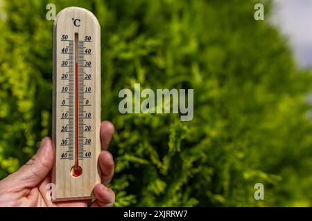 Un uomo tiene in mano un termometro di legno in giardino che indica una temperatura molto elevata - protezione contro il calore Foto Stock