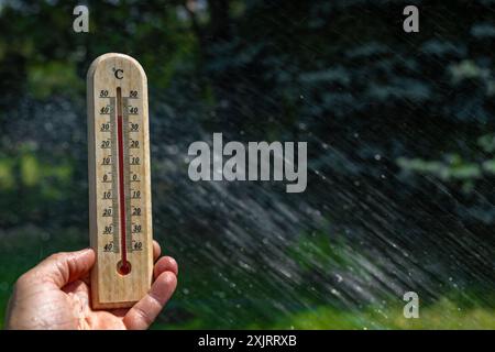 Un uomo tiene in mano un termometro di legno in giardino che indica una temperatura molto elevata - protezione contro il calore Foto Stock